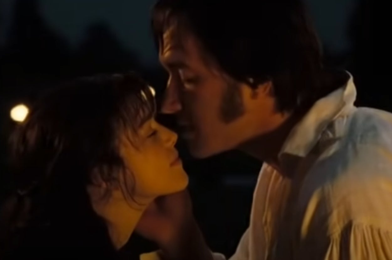 Mr. Darcy and Elizabeth in "Pride and Prejudice" in period attire share an intimate moment, gently leaning in for a kiss with closed eyes in a dimly lit setting