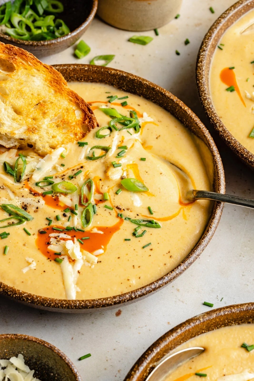 A bowl of creamy soup topped with sliced green onions, shredded cheese, and a drizzle of hot sauce, accompanied by a piece of toasted bread