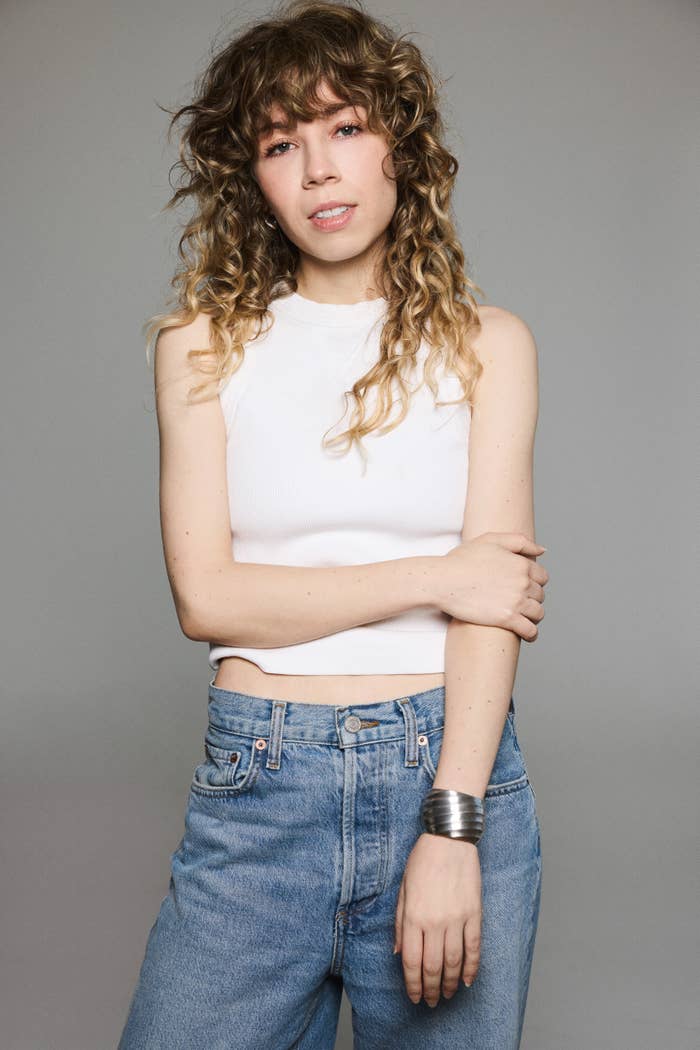 Person in casual outfit with curly hair, wearing a sleeveless top and jeans, posing with a neutral expression