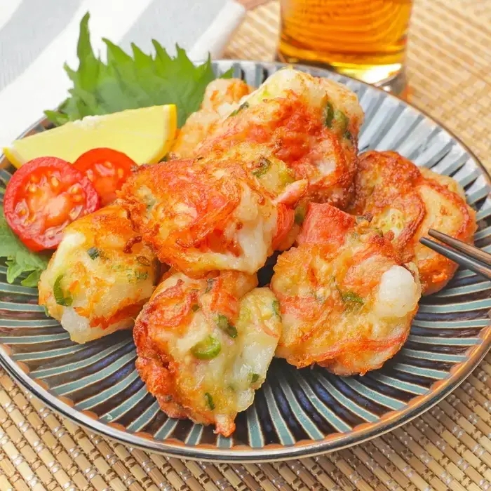 皿に盛られた豆腐と餅の落とし揚げ、レモン、トマトのスライス、しそが添えられ、飲み物が背景にある。