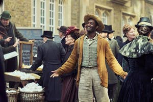 Actor in period costume smiles amid Victorian street fair, surrounded by people in historical attire