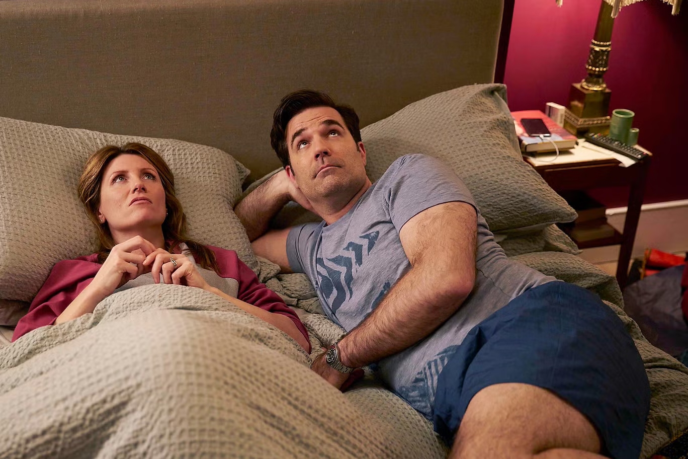 Two people lying in bed looking contemplative, the woman on the left holds a pillow and the man on the right has one arm behind his head