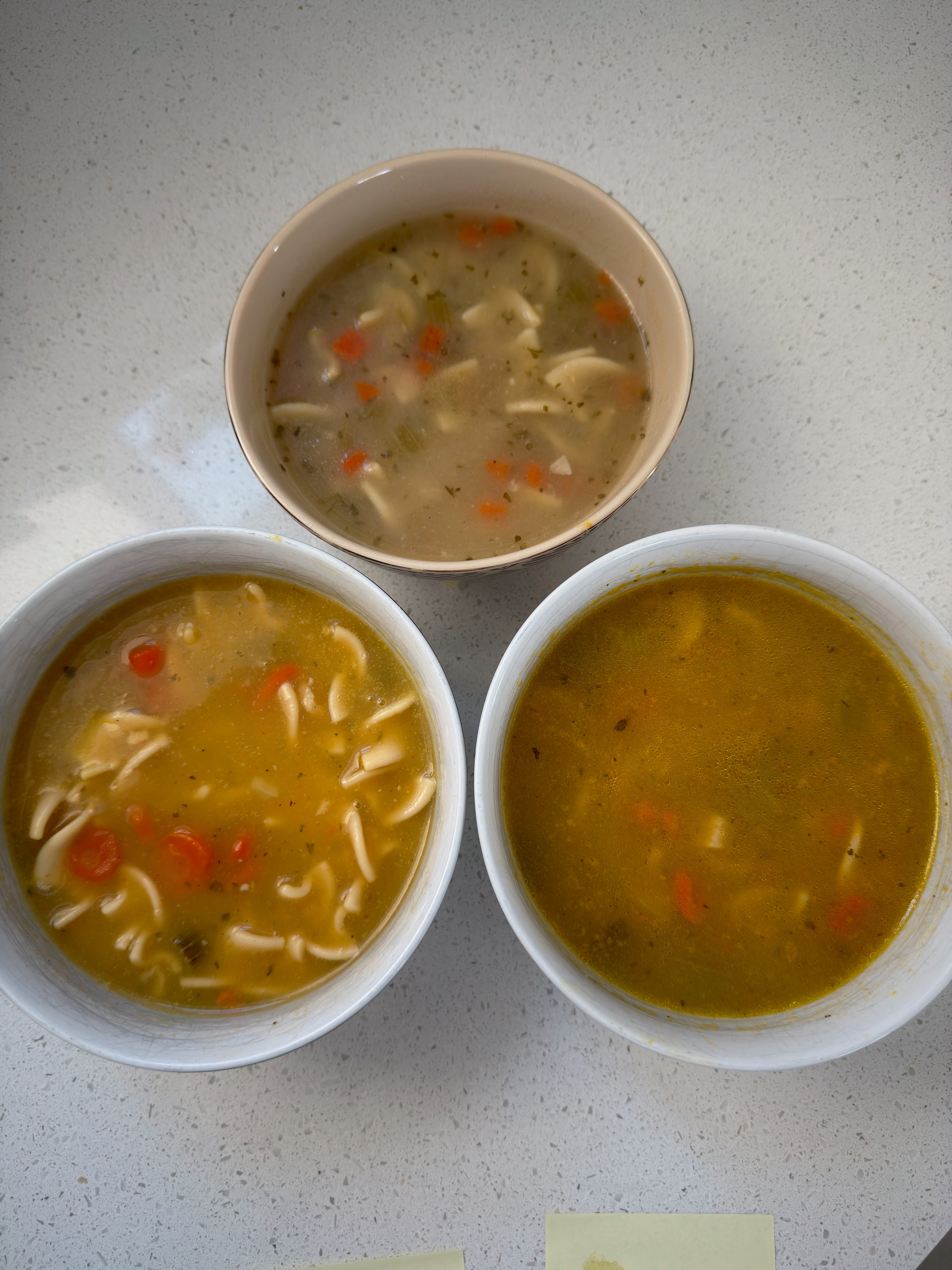Three bowls of soup with visible ingredients like noodles and vegetables, arranged on a white surface, showcasing different textures