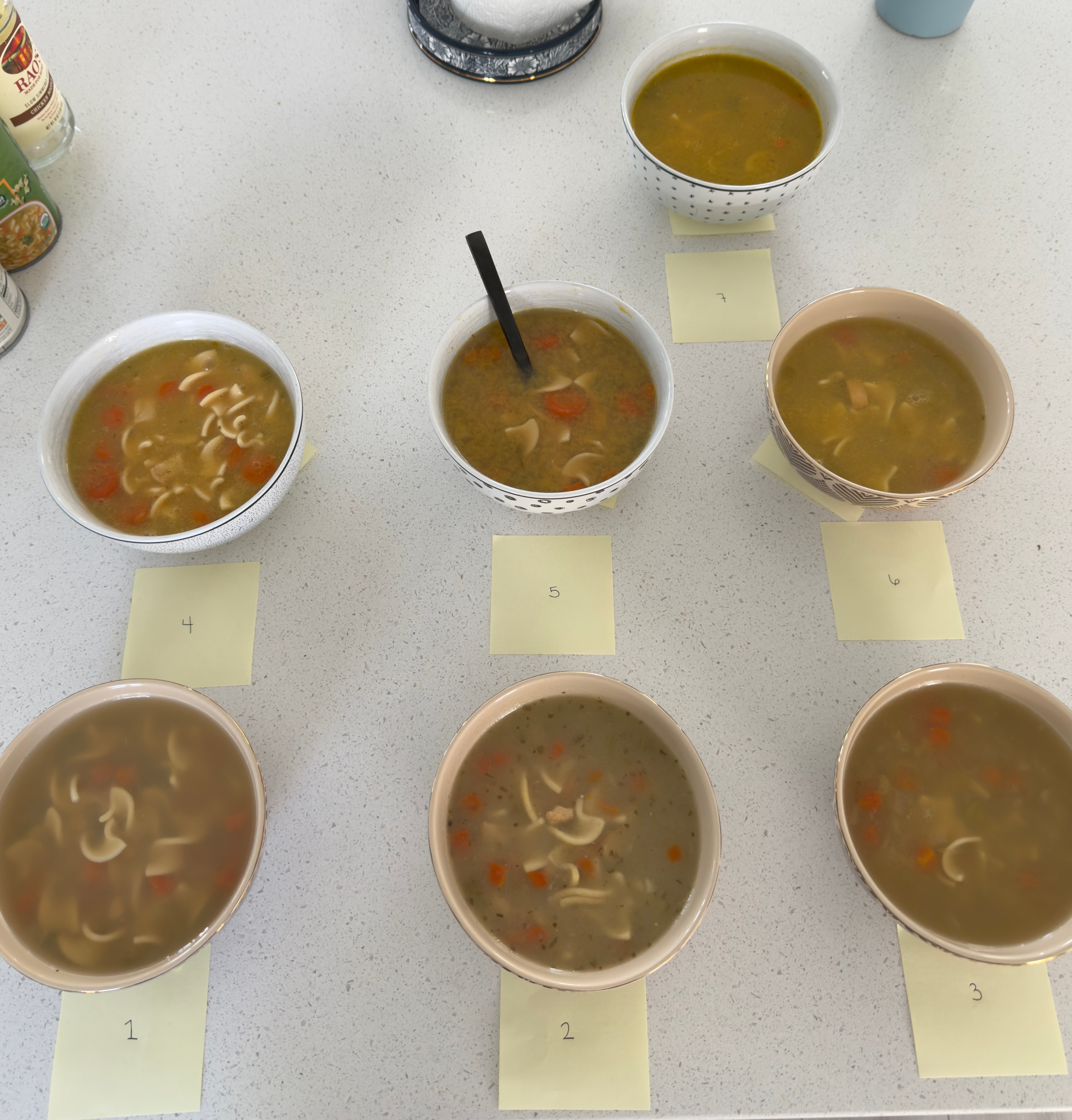 Seven bowls of soup with various ingredients, each numbered 1 to 7 on sticky notes, arranged on a countertop