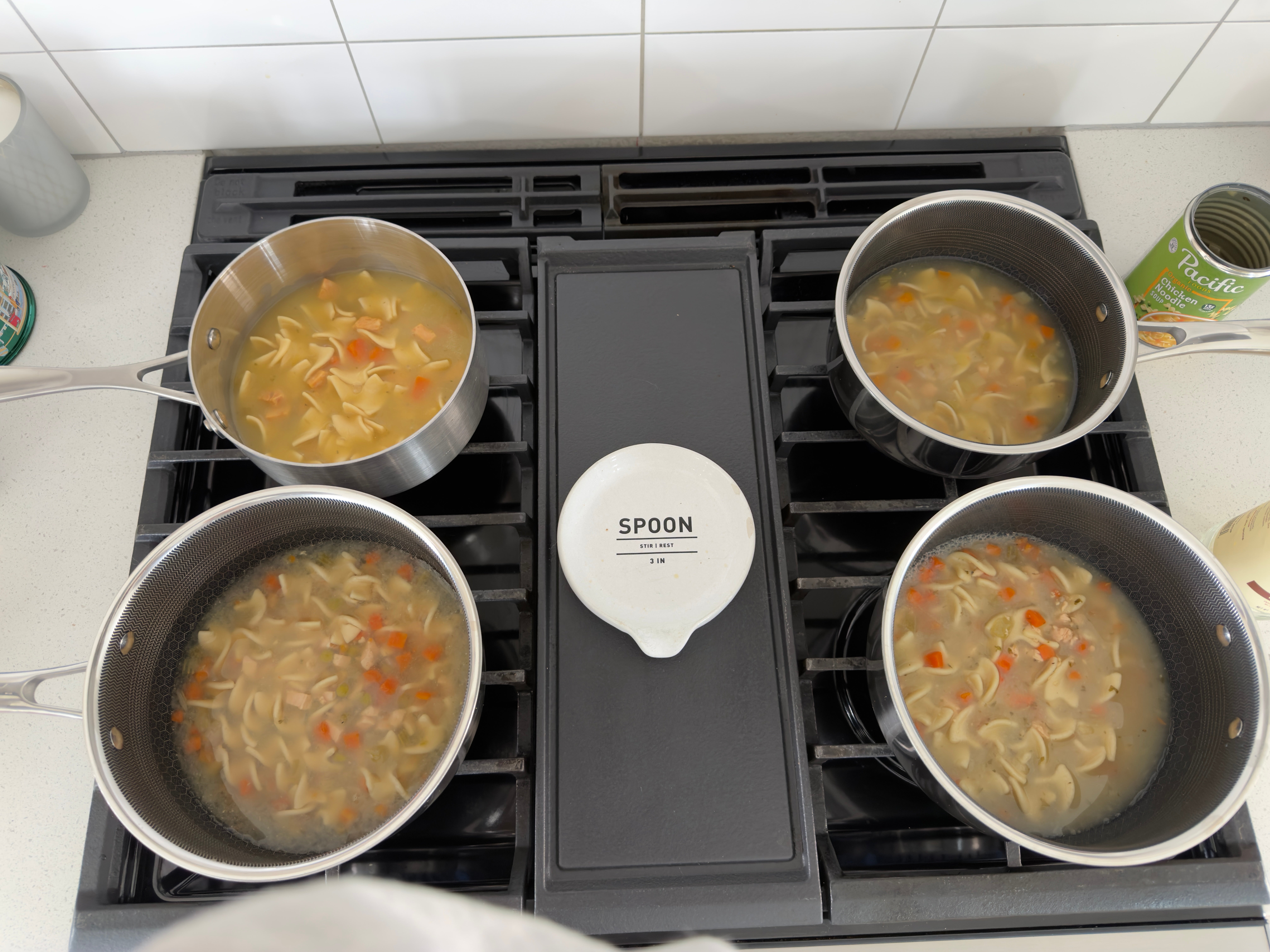 Four pots on a stovetop filled with noodle soup, containing visible vegetables. A can of peas is nearby