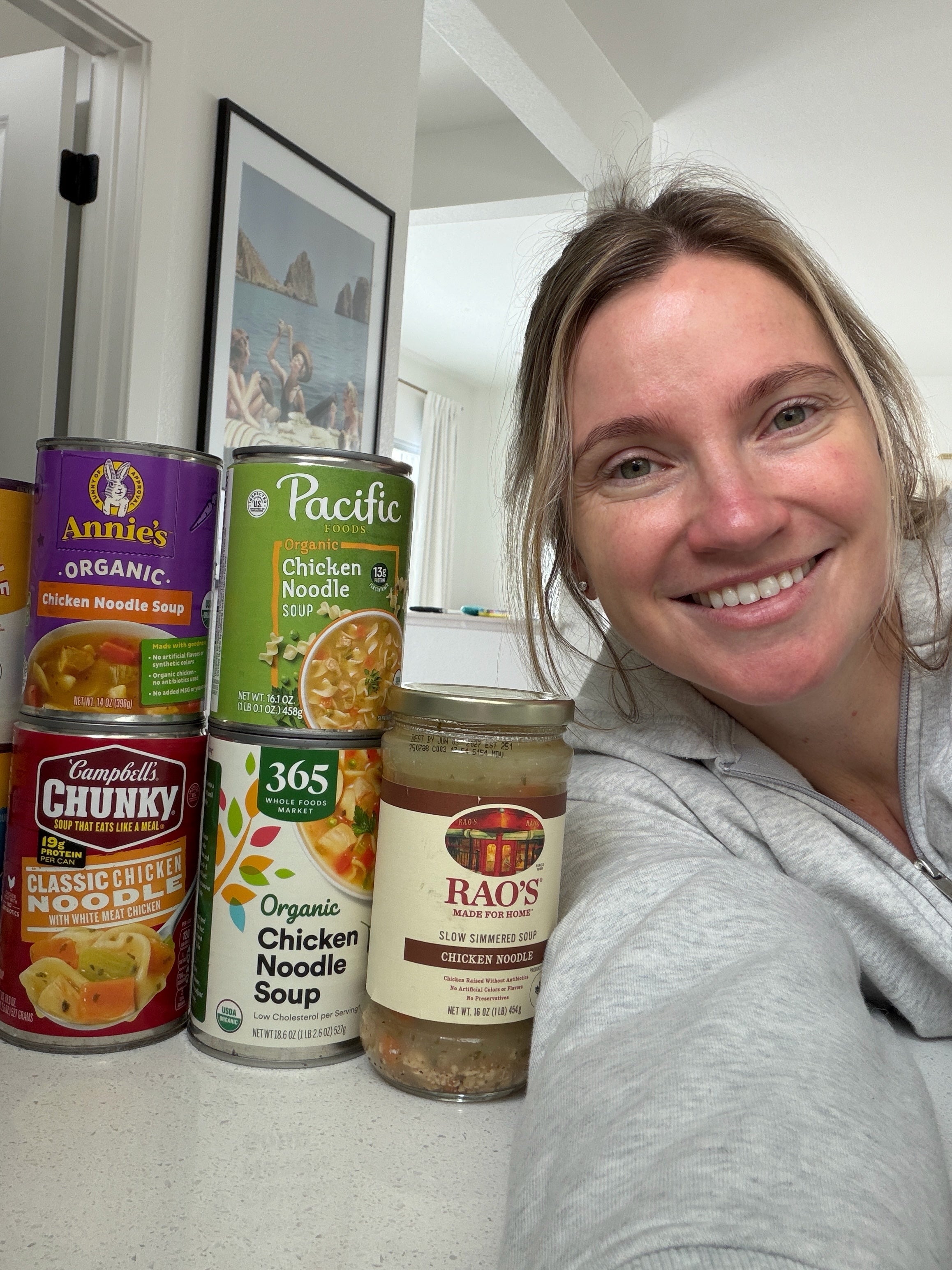 Woman smiling next to a stack of different chicken noodle soup cans and a jar of sauce on a kitchen counter