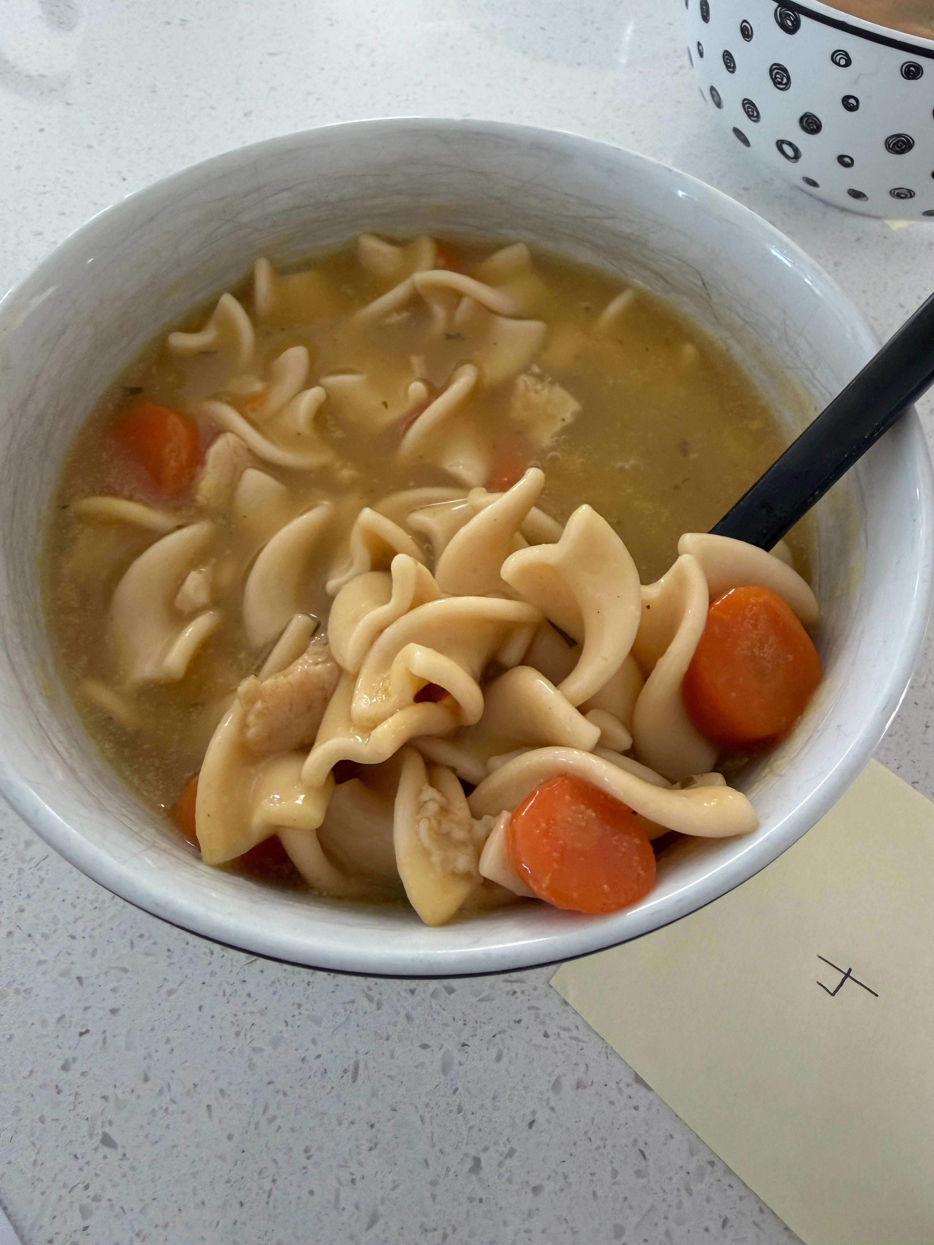 A bowl of noodle soup with carrots and a spoon on a countertop