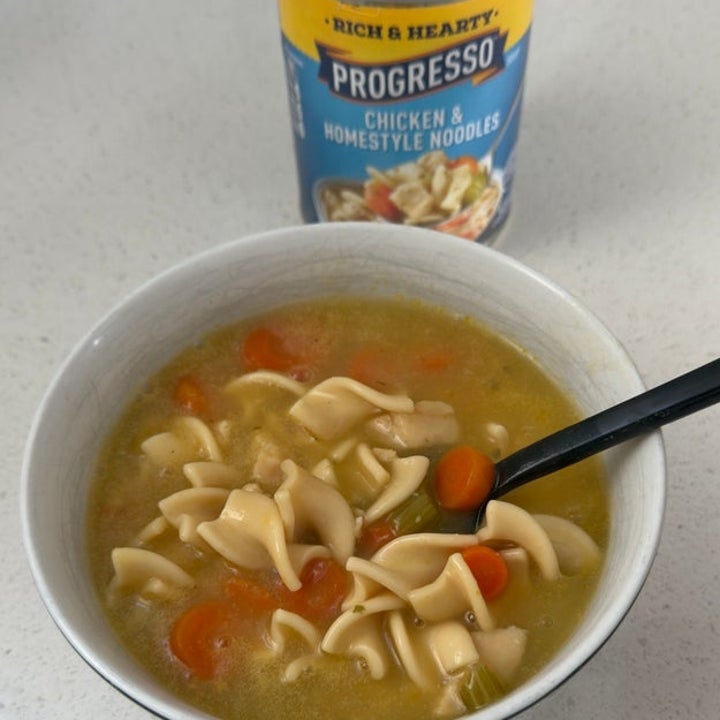 A bowl of chicken noodle soup with a spoon, next to an open can of Progresso Chicken &amp; Homestyle Noodles on a countertop