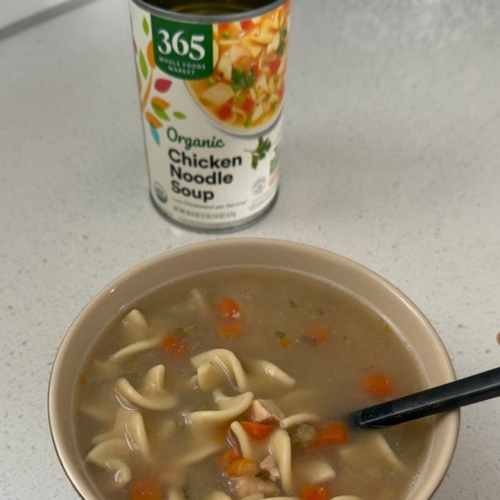 A bowl of chicken noodle soup with vegetables sits on a counter, next to an open can of 365 Organic Chicken Noodle Soup