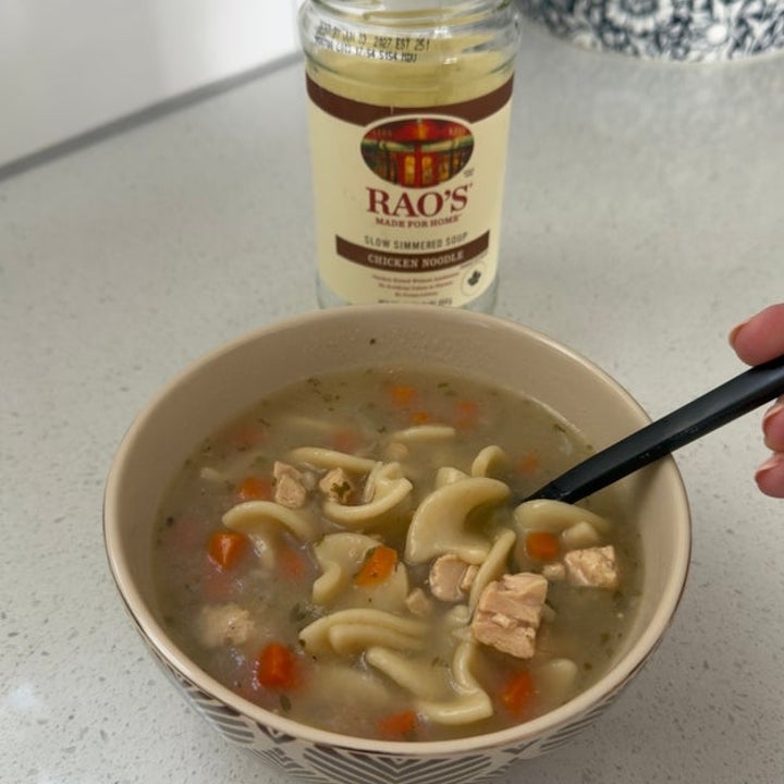 A bowl of chicken noodle soup with a spoon, featuring pieces of chicken, carrots, and noodles. A jar of chicken broth is in the background