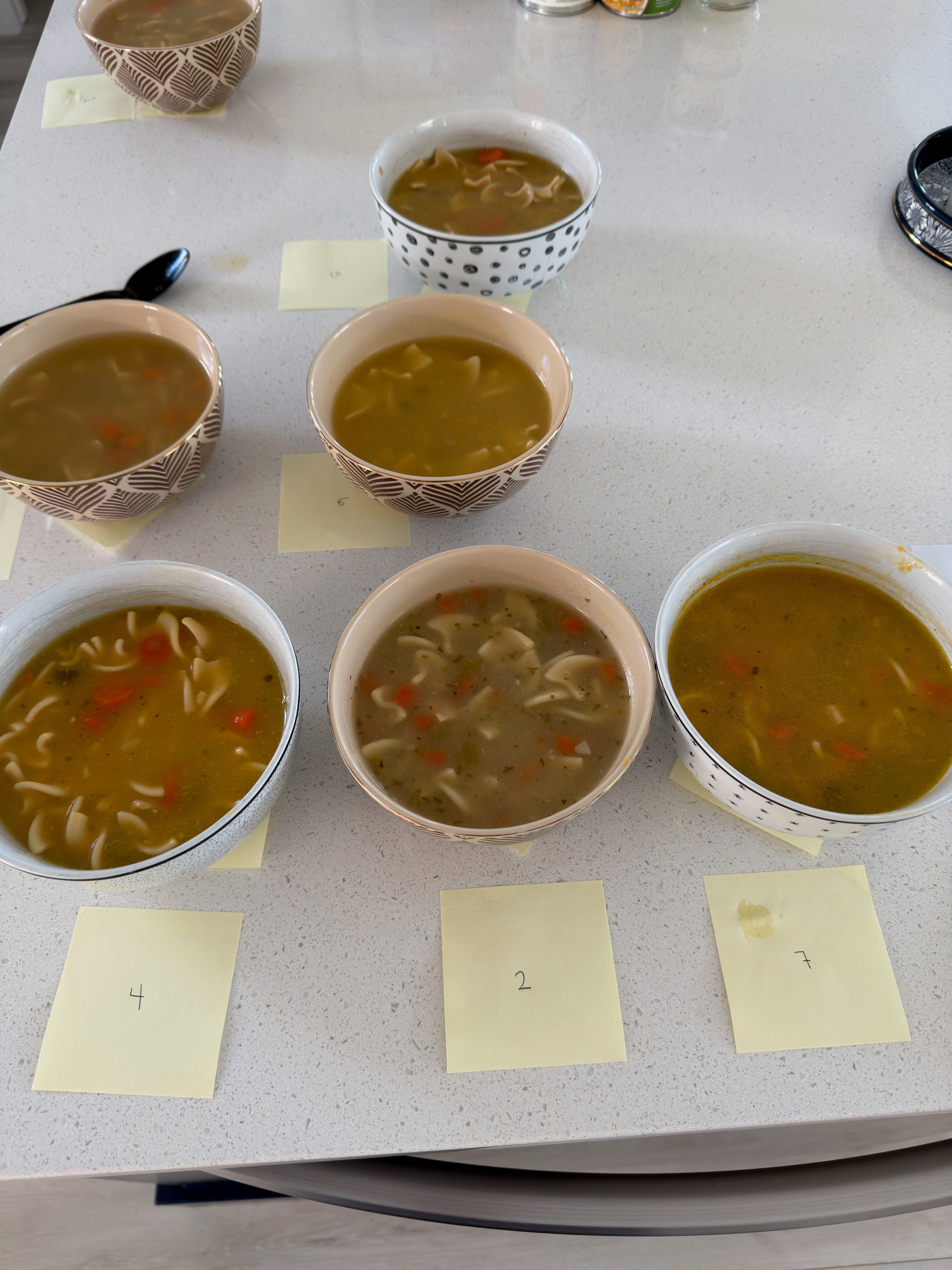 Five bowls of noodle soup with carrots on a counter, each labeled with numbered sticky notes for comparison
