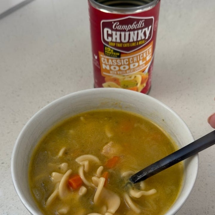 A bowl of noodle soup with vegetables and a spoon, next to an opened can of Campbell's Chunky Classic Chicken Noodle Soup