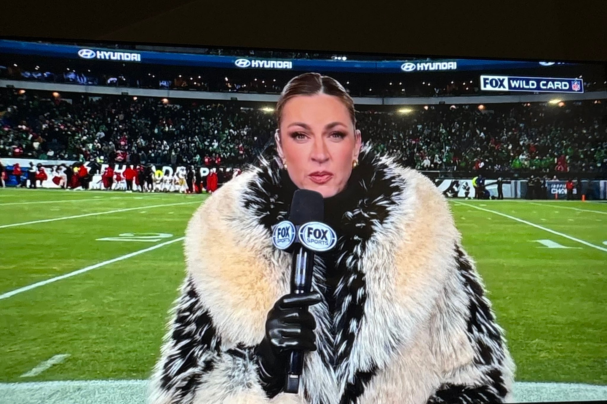 大きなスタジアムで、マイクを持ち、豪華なファーコートを着たリポーターがカメラに向かっているシーン。