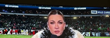 大きなスタジアムで、マイクを持ち、豪華なファーコートを着たリポーターがカメラに向かっているシーン。