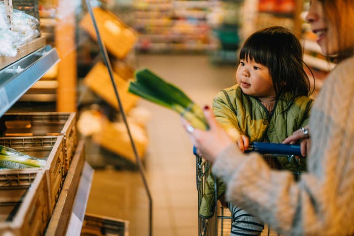 Child sitting in shopping cart inside a supermarket, looking at fresh produce while an adult holds vegetables