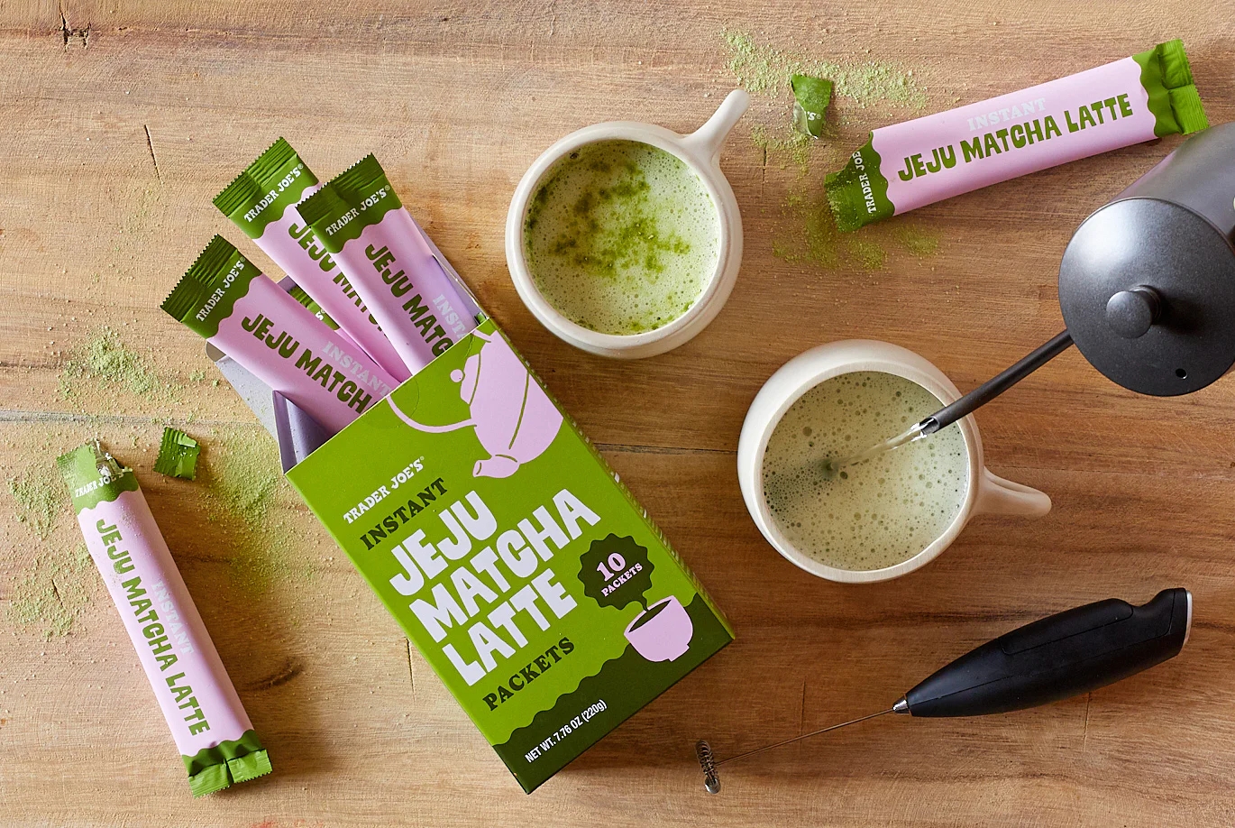 Box of instant Jeju matcha latte packets on a wooden surface with brewed matcha lattes in mugs and a whisk nearby