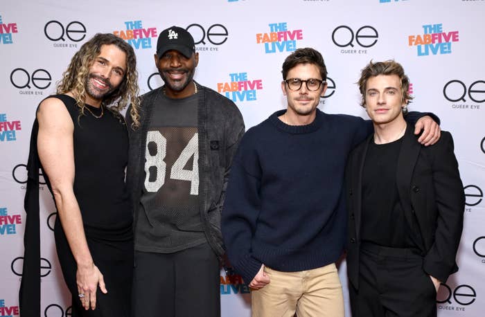 Group of four people at a "Queer Eye" event; casual and semi-formal attire, posing in front of a step-and-repeat with show logos