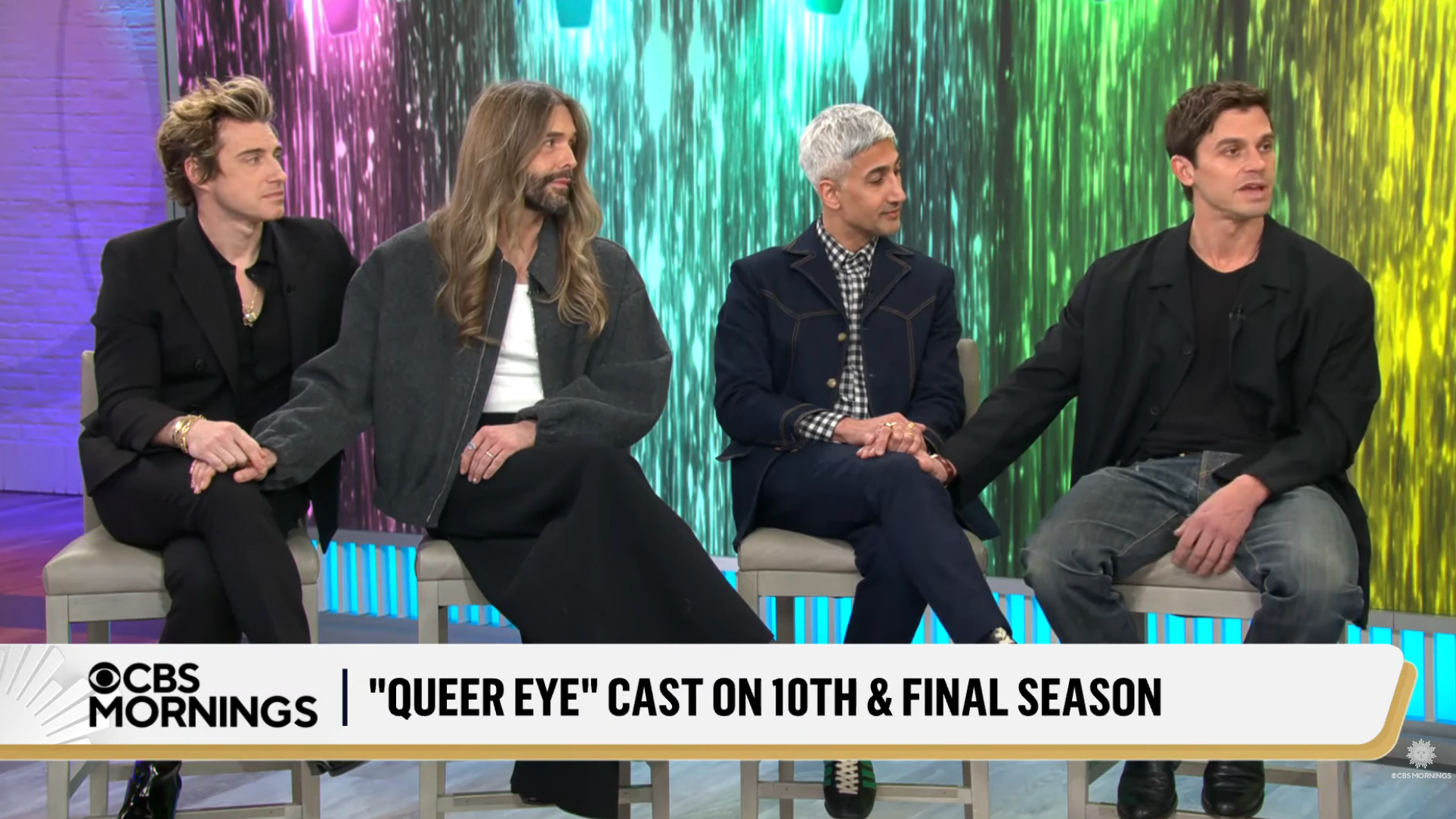 Four cast members sit on a TV set for a "Queer Eye" interview about the 10th and final season, holding hands, with a colorful background