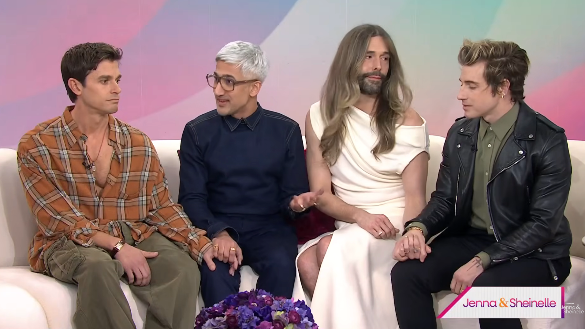 Group of people sitting on a talk show set, engaging in conversation, with one in a white dress and another in a leather jacket