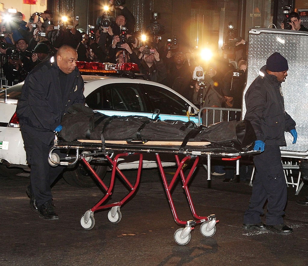 To personer i uniformer skyver en båre som holder en tildekket figur. Kameraer blinker mens fotografer fanger scenen, noe som indikerer en høyprofilert begivenhet