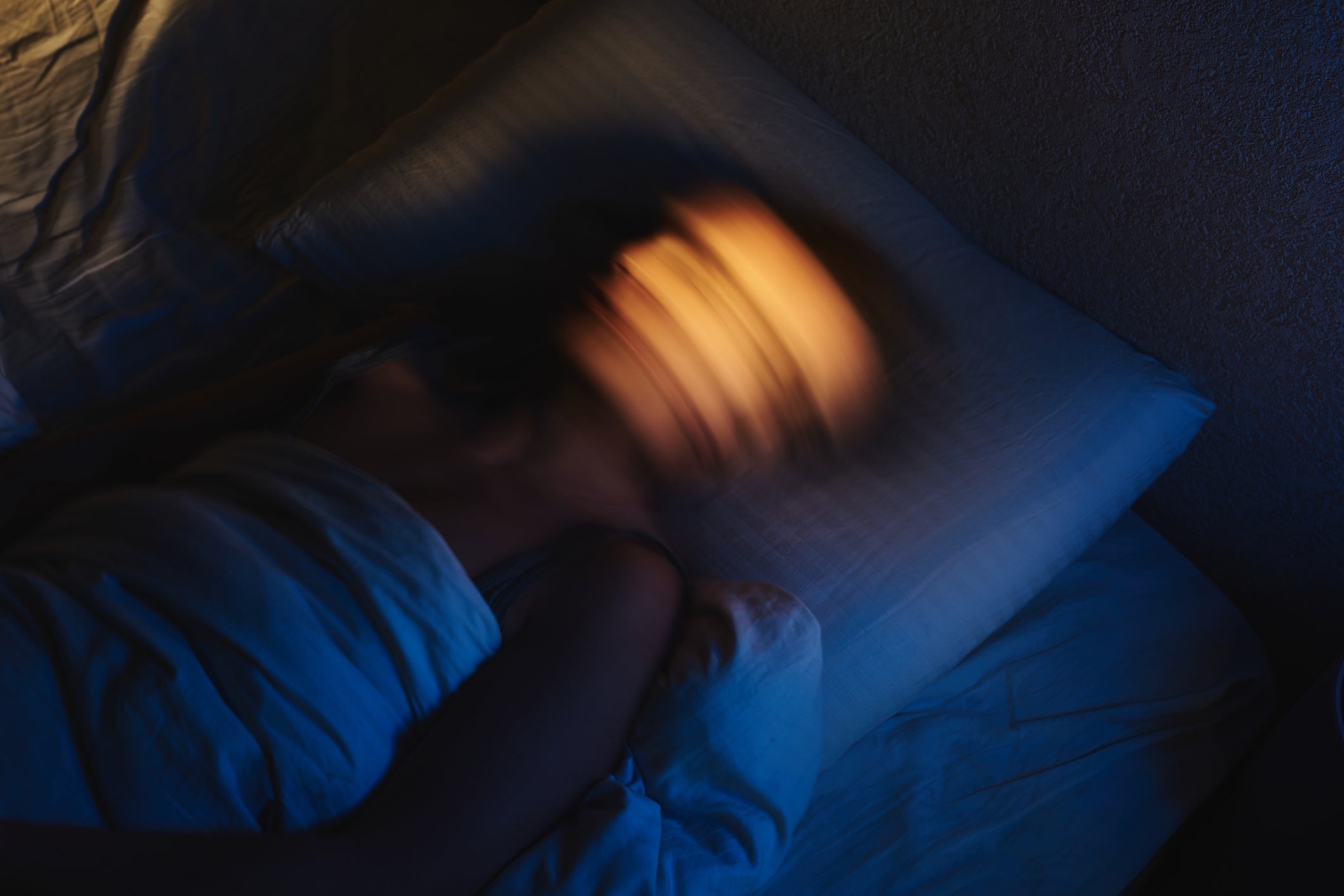 Person i sengen med et uskarpt ansikt, noe som tyder på bevegelse eller rastløshet, under tepper i et svakt opplyst rom