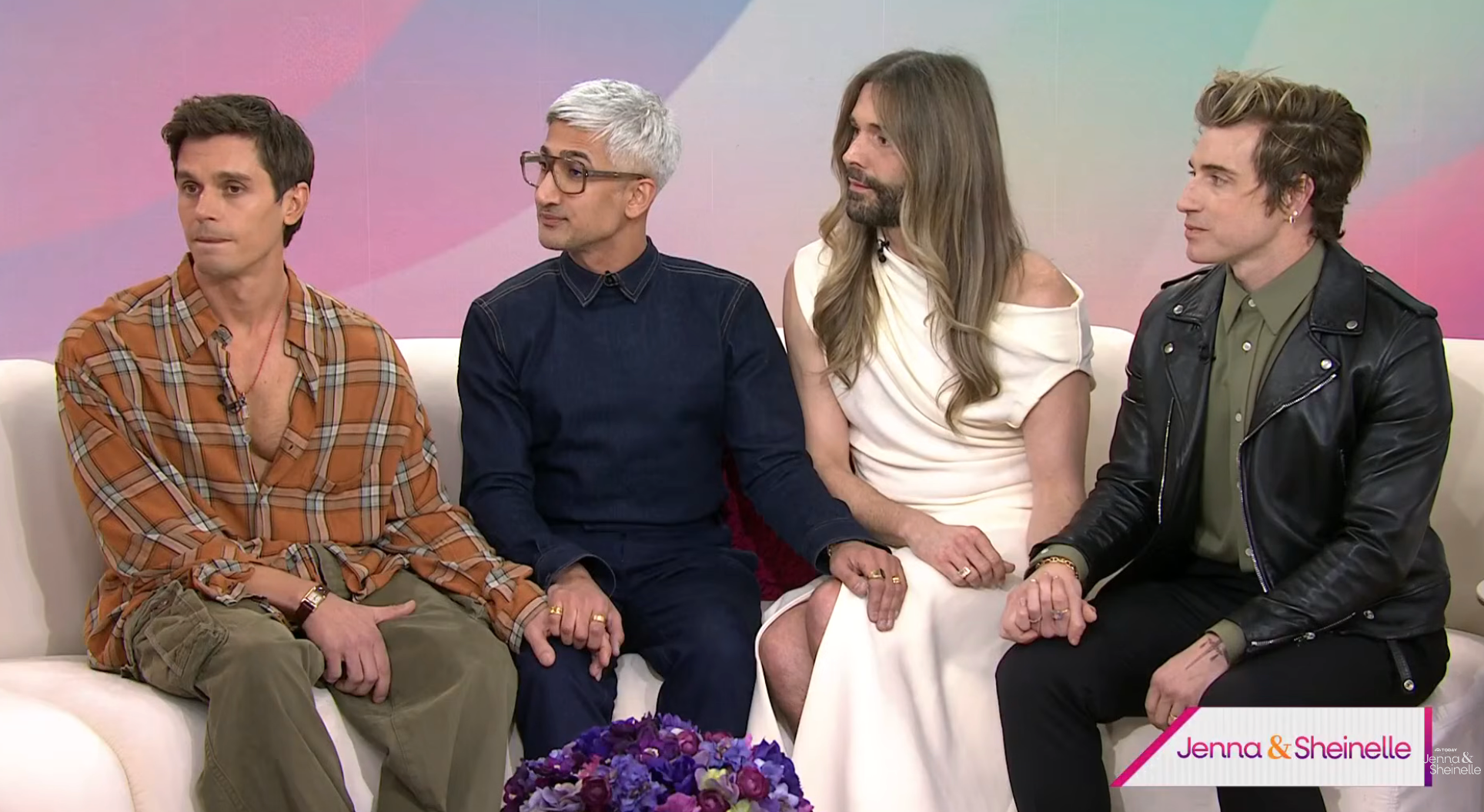 Four people sit together on a sofa. Two wear casual outfits, one a dark formal look, and another a white dress. They appear focused and attentive