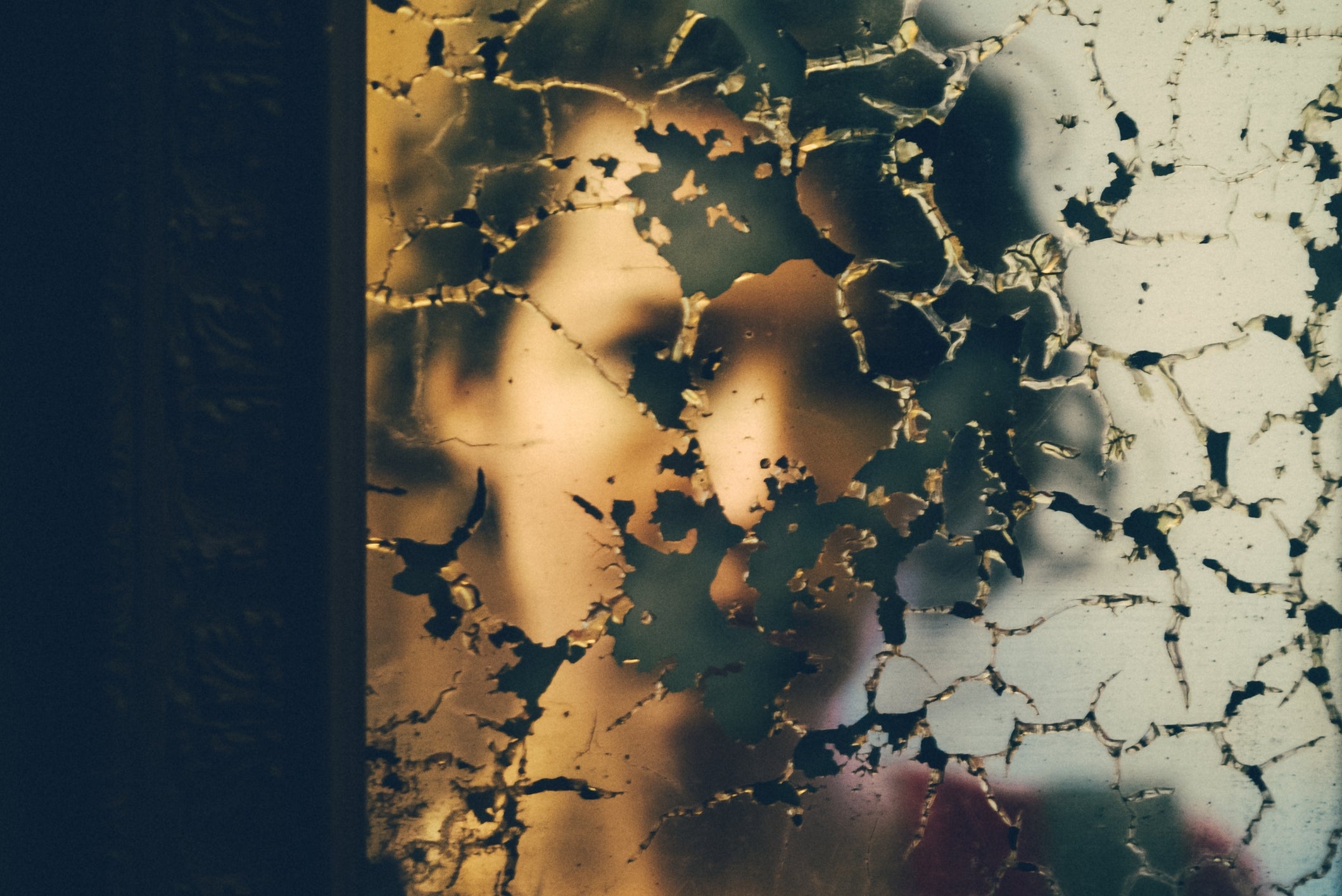 En uskarp refleksjon av en person bak et sprukket speil i vintagestil som skaper en abstrakt, mystisk effekt