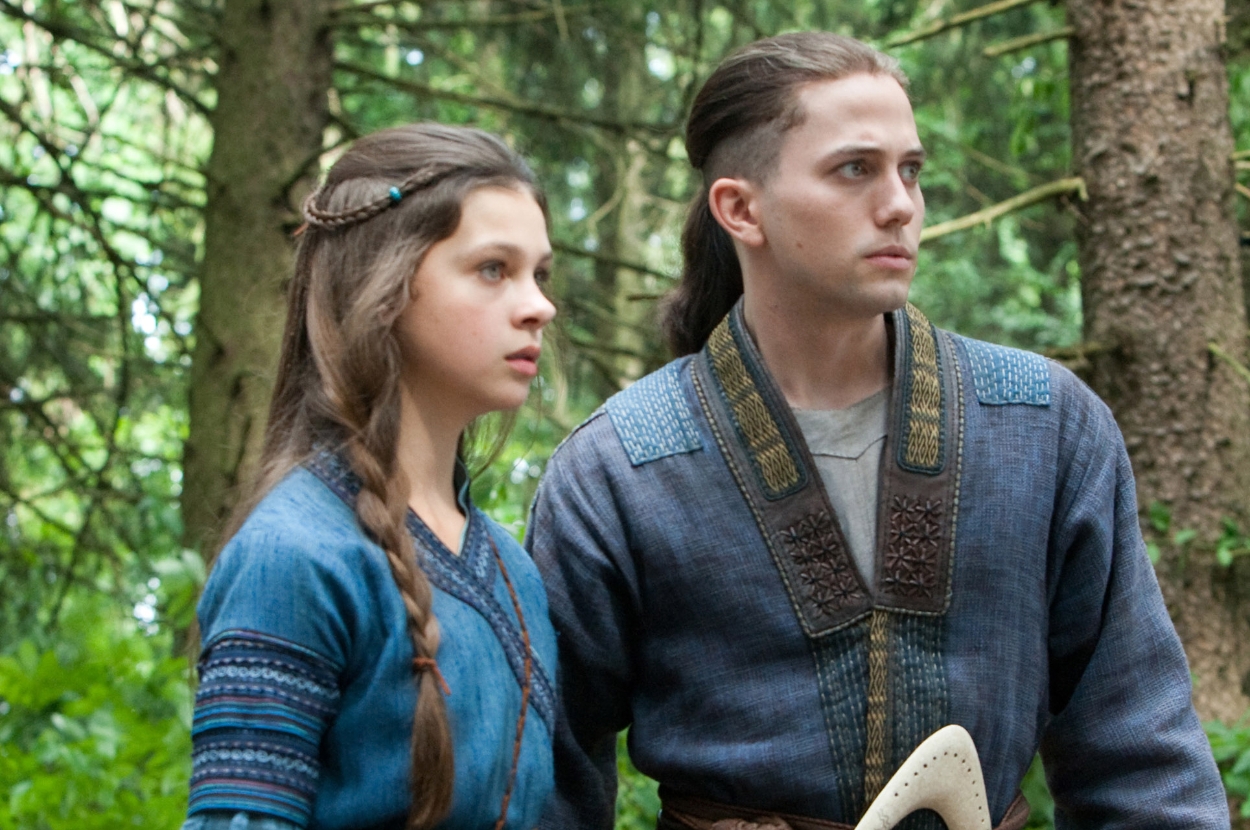 Two individuals stand in a forest setting, dressed in elaborate, medieval-style costumes, looking intently into the distance