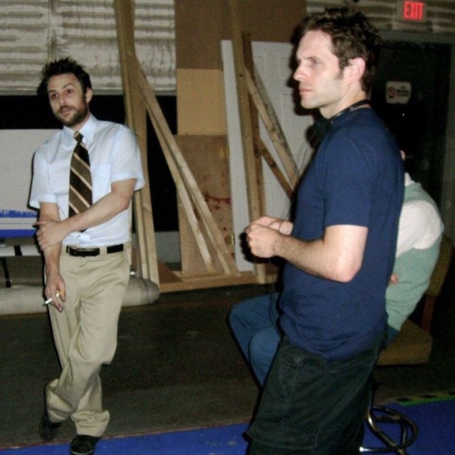 Two men basal   casually successful  a country   with woody  panels; 1  wears a necktie  and holds a cigarette, portion    the different   is dressed successful  a T-shirt and jeans