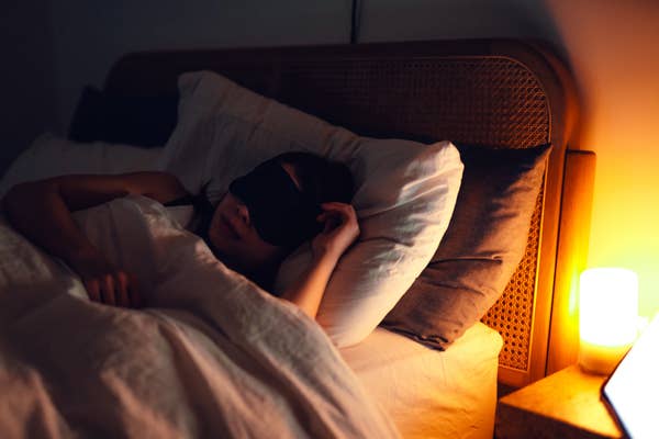 Person sleeping in bed with an eye mask, next to a softly lit bedside lamp, creating a calm and restful atmosphere