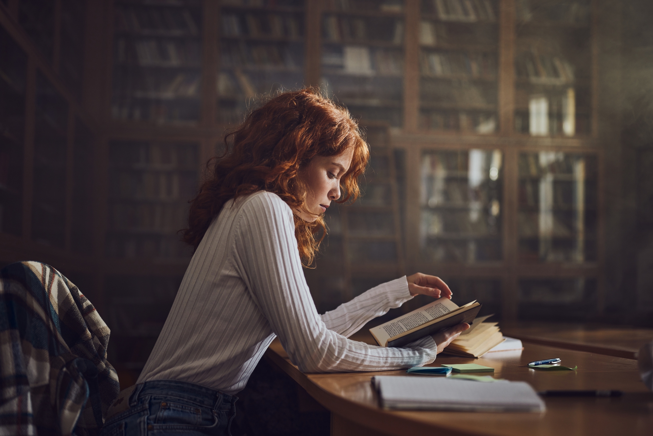 A person with curly hair reads a book in a library, seated at a desk with papers and a pen nearby