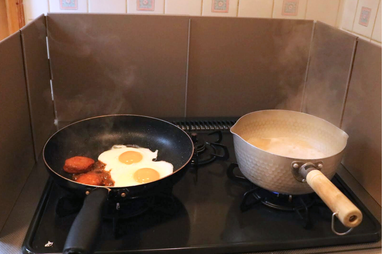 フライパンで目玉焼きとソーセージを調理中、隣で鍋で何かが沸騰しているキッチンの様子。