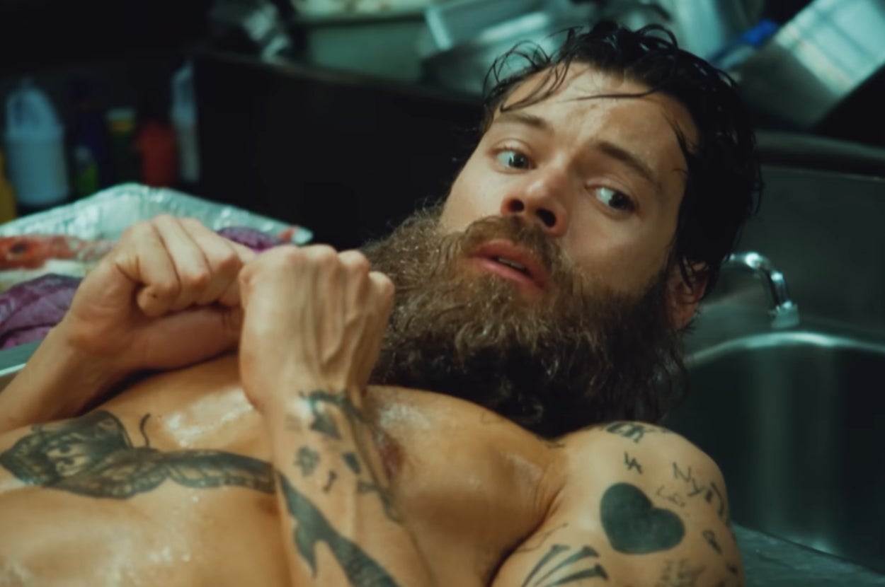 A man with tattoos, wet hair, and a beard appears surprised while lying in a metal tub surrounded by kitchen items
