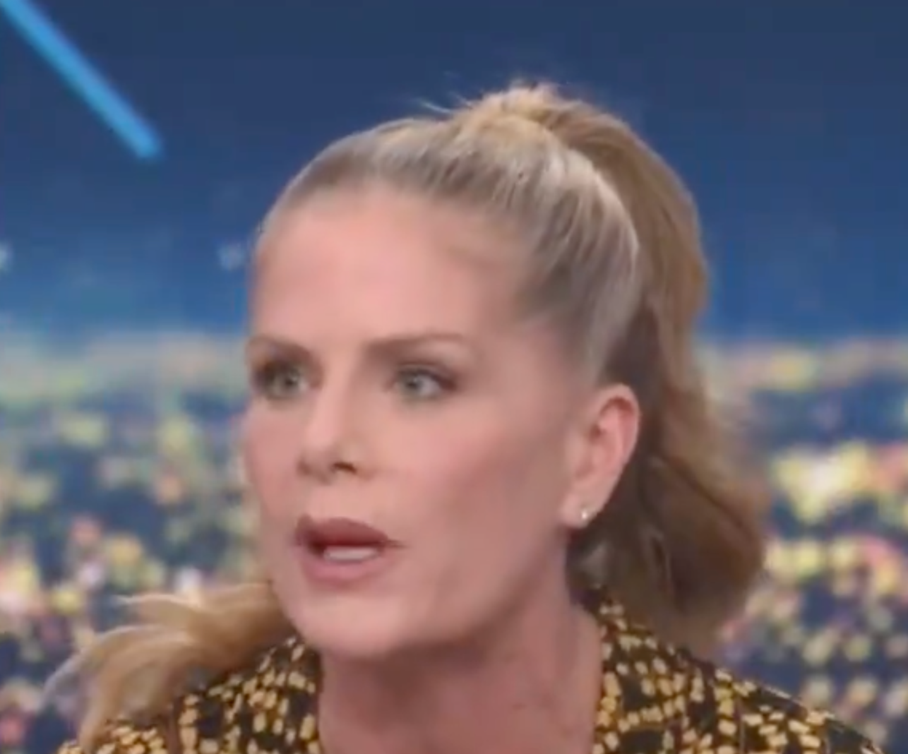 A person is speaking during a news segment, wearing a patterned top and with their hair in a ponytail