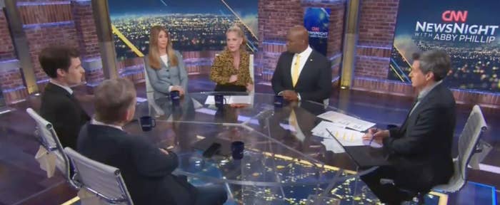 Panel discussion on set with six people around a glass table on a news show. Cityscape visible in the background