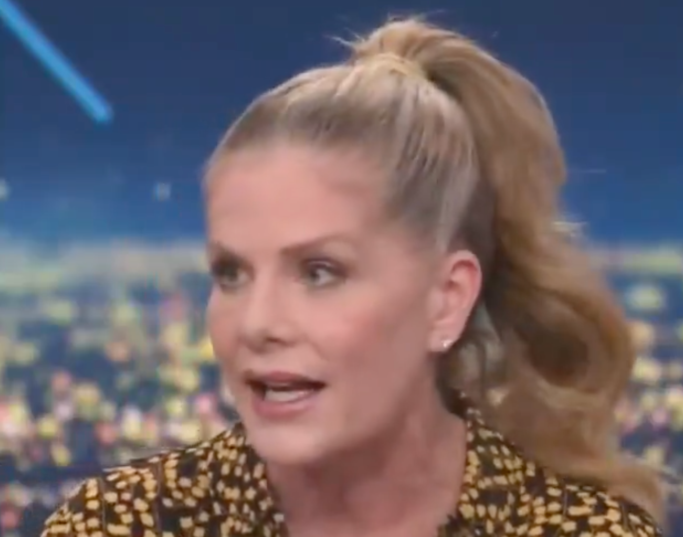 Person with a ponytail speaking on a broadcast set, wearing a patterned blouse