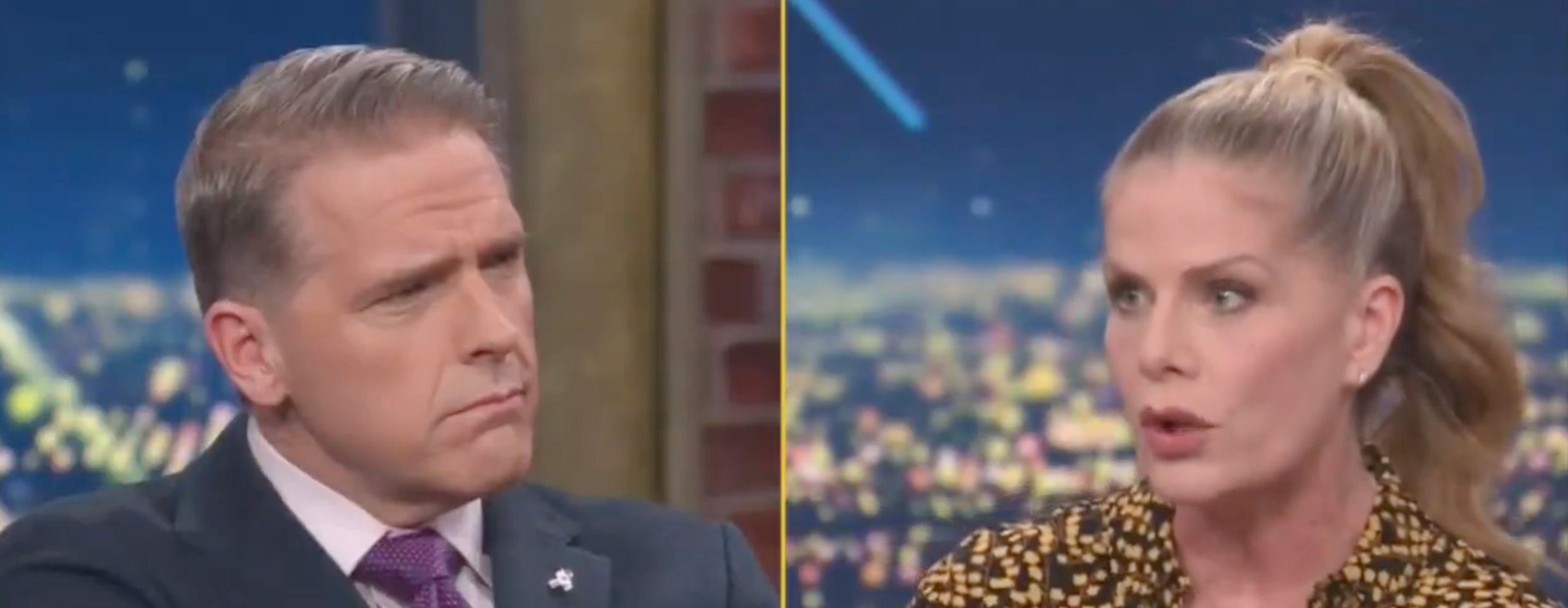 Two people are engaged in a serious discussion on a TV talk show set, with cityscape visible in the background