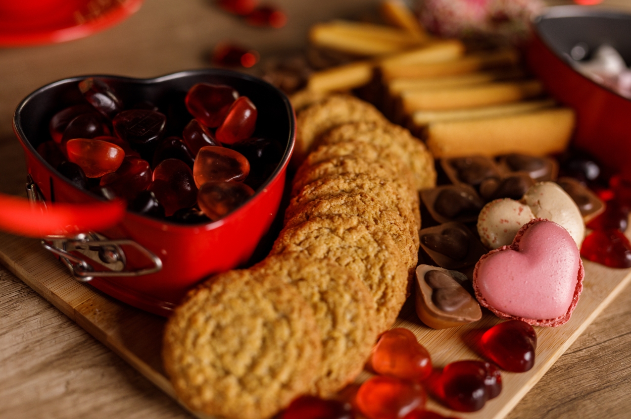 Assorted heart-shaped candies and cookies on a wooden board, featuring chocolates and a heart-shaped tin full of gummy hearts
