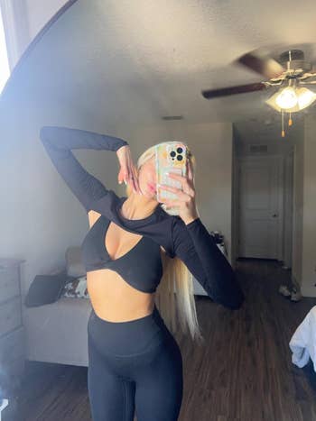 Person taking a mirror selfie, wearing a stylish black bolero, sports bra, and leggings in a bedroom setting