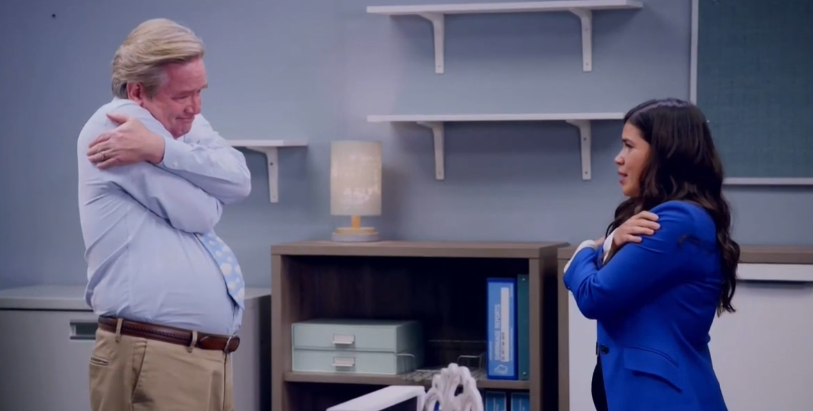 Two people in an office, standing and hugging themselves, one wearing a blue blazer