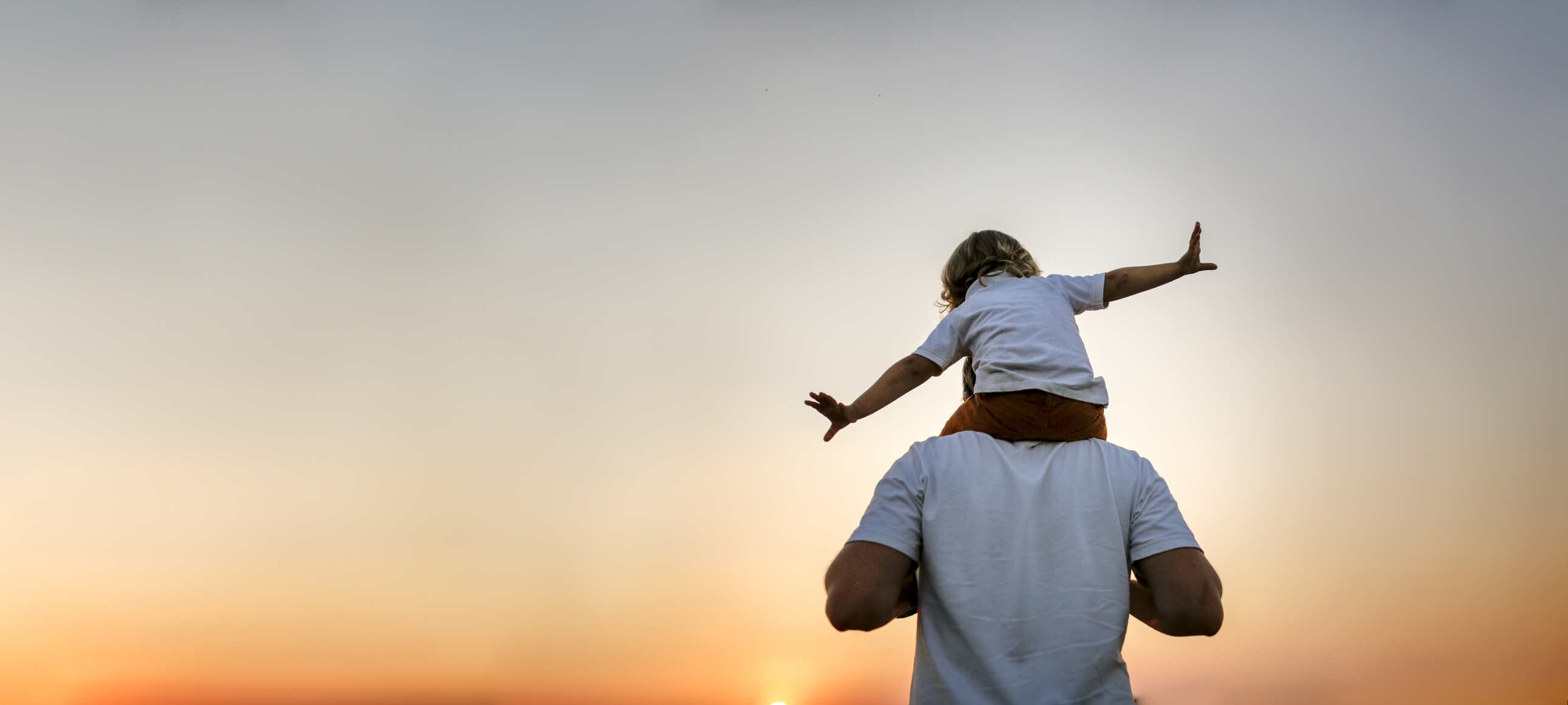 A child sits on an adult's shoulders, arms outstretched, against a sunset sky