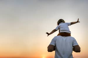 A child sits on an adult's shoulders, arms outstretched, against a sunset sky