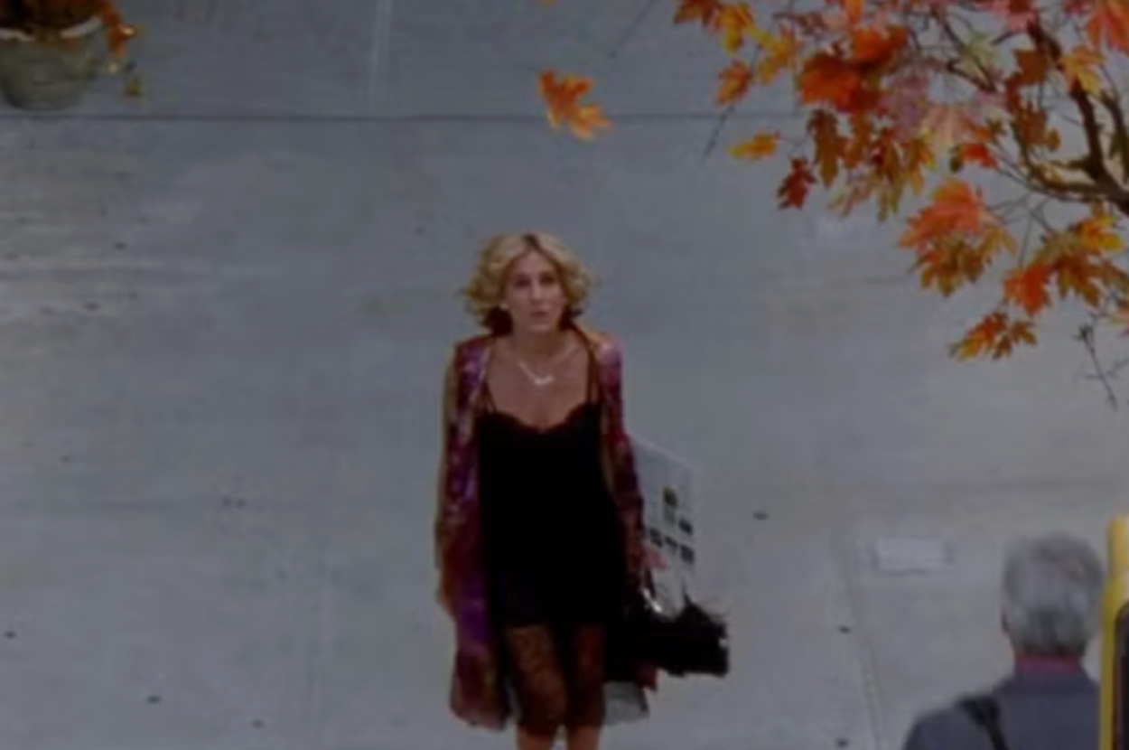 Person with curly hair walking on a sidewalk, wearing a stylish black dress and patterned coat, carrying a shopping bag. Autumn leaves visible