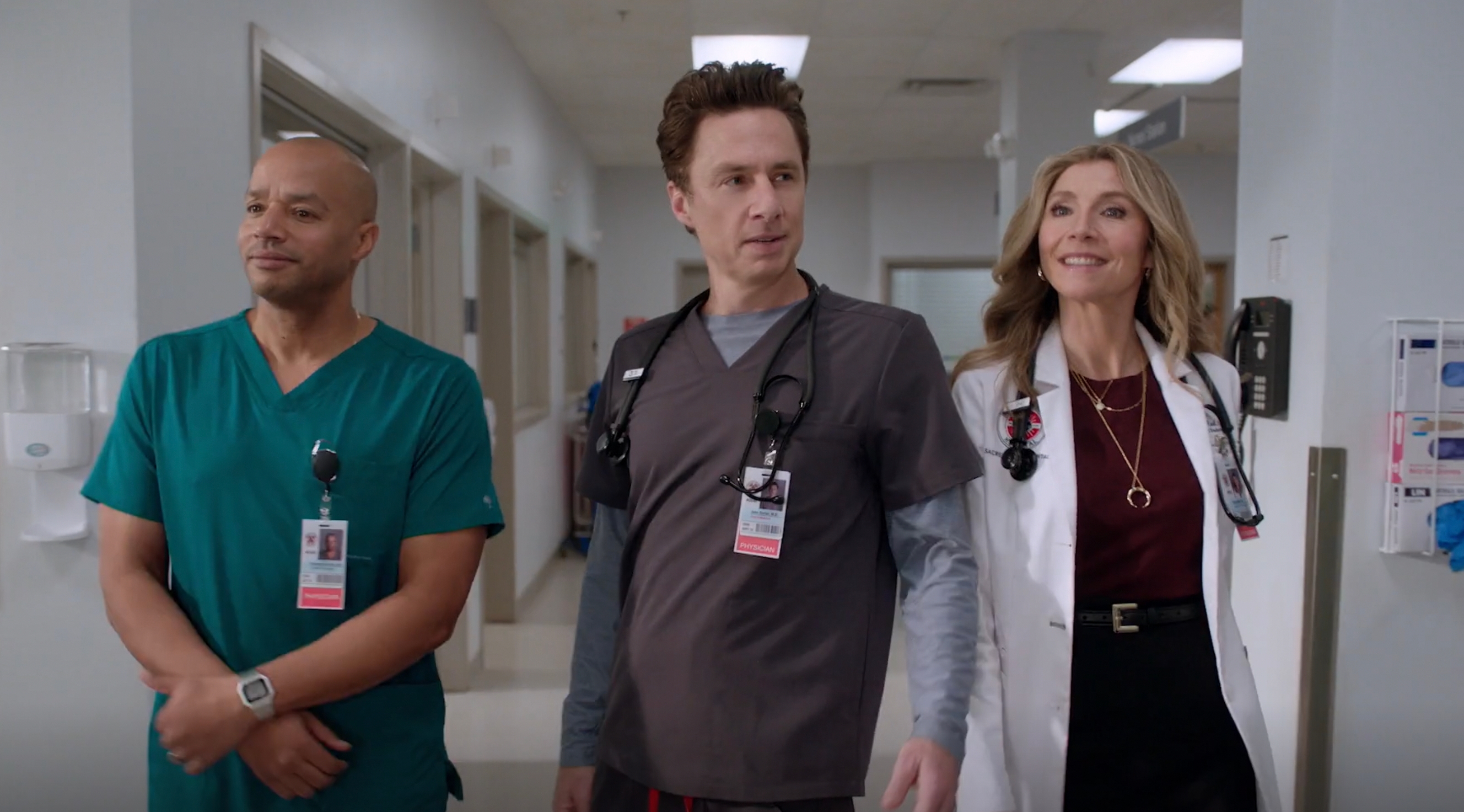 Three actors in medical attire walk down a hospital corridor, each wearing scrubs and stethoscopes, suggesting a scene from a medical TV show