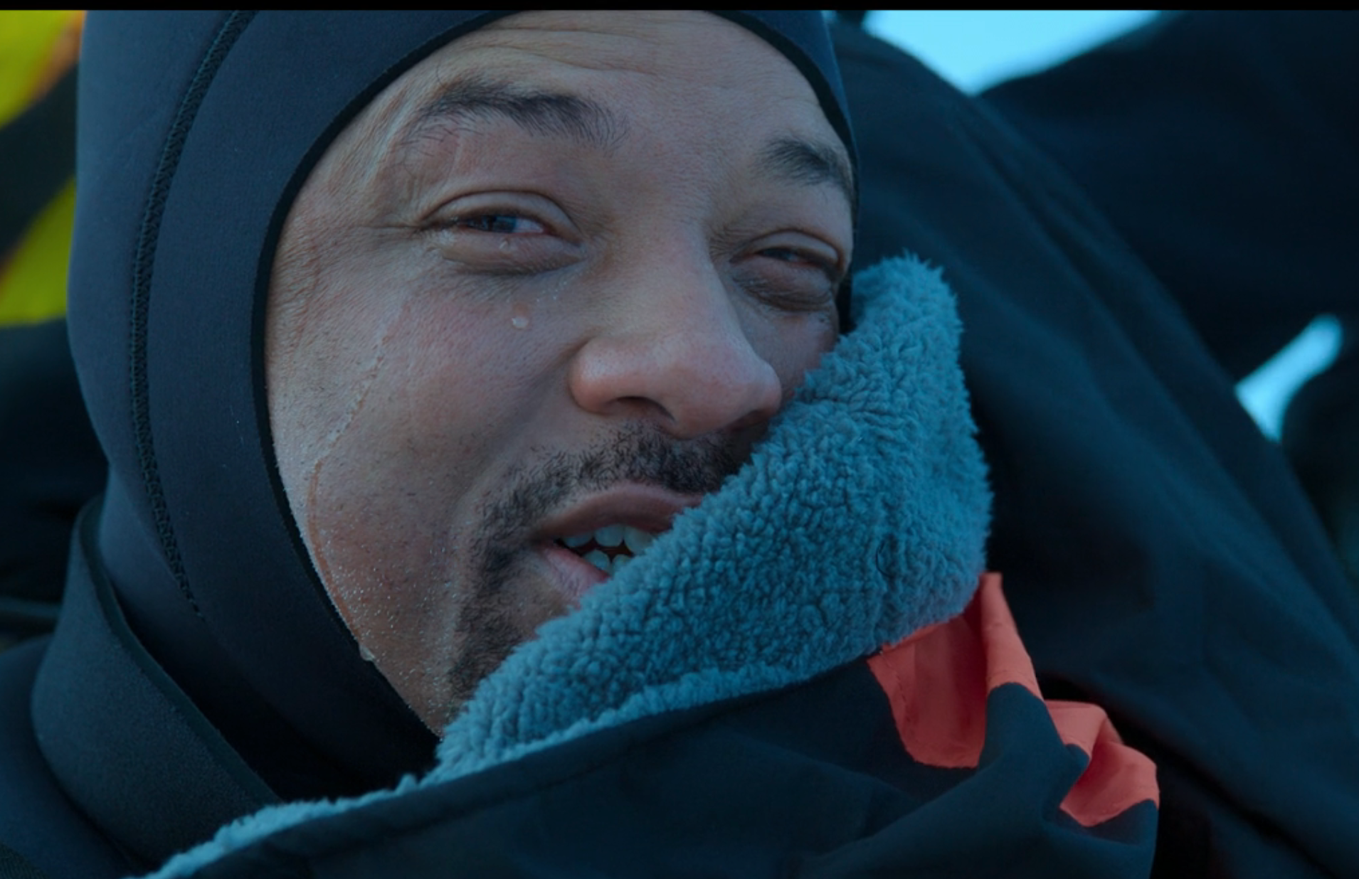 A person in a black hood, bundled in a fleece jacket outdoors, with a joyful smile and teary eyes