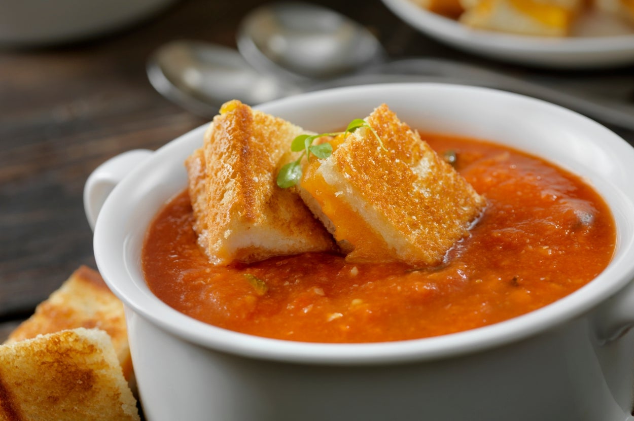 Tomato soup topped with crispy grilled cheese croutons in a white bowl, with silver spoons in the background