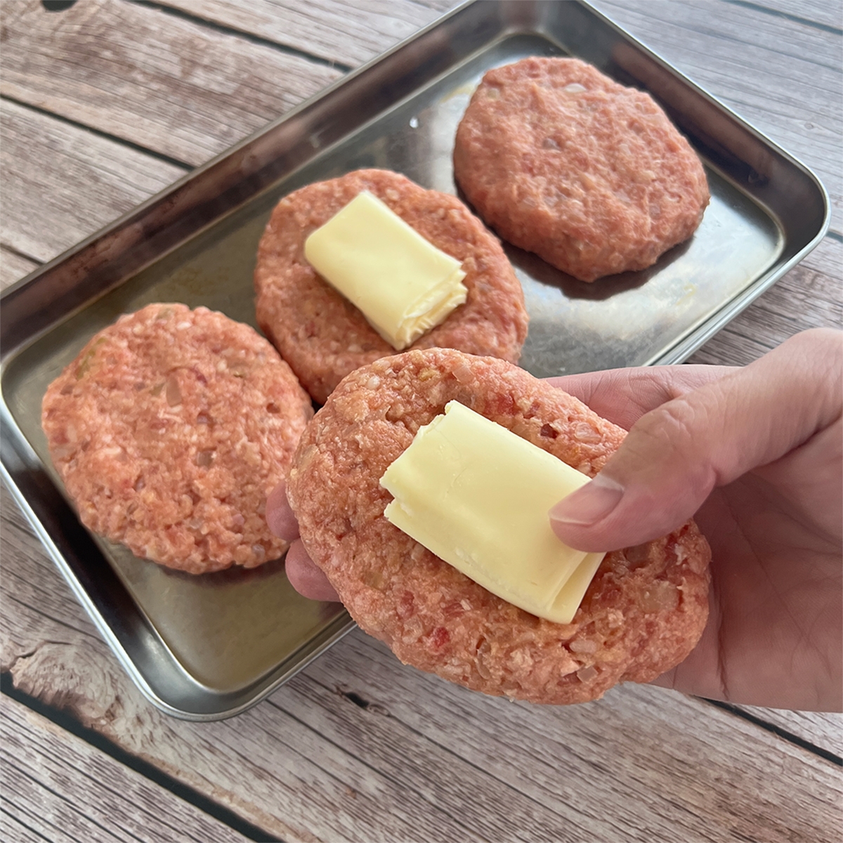 ハンバーグパティにチーズをのせて手に持っている様子。金属トレーに他のパティも準備されている。