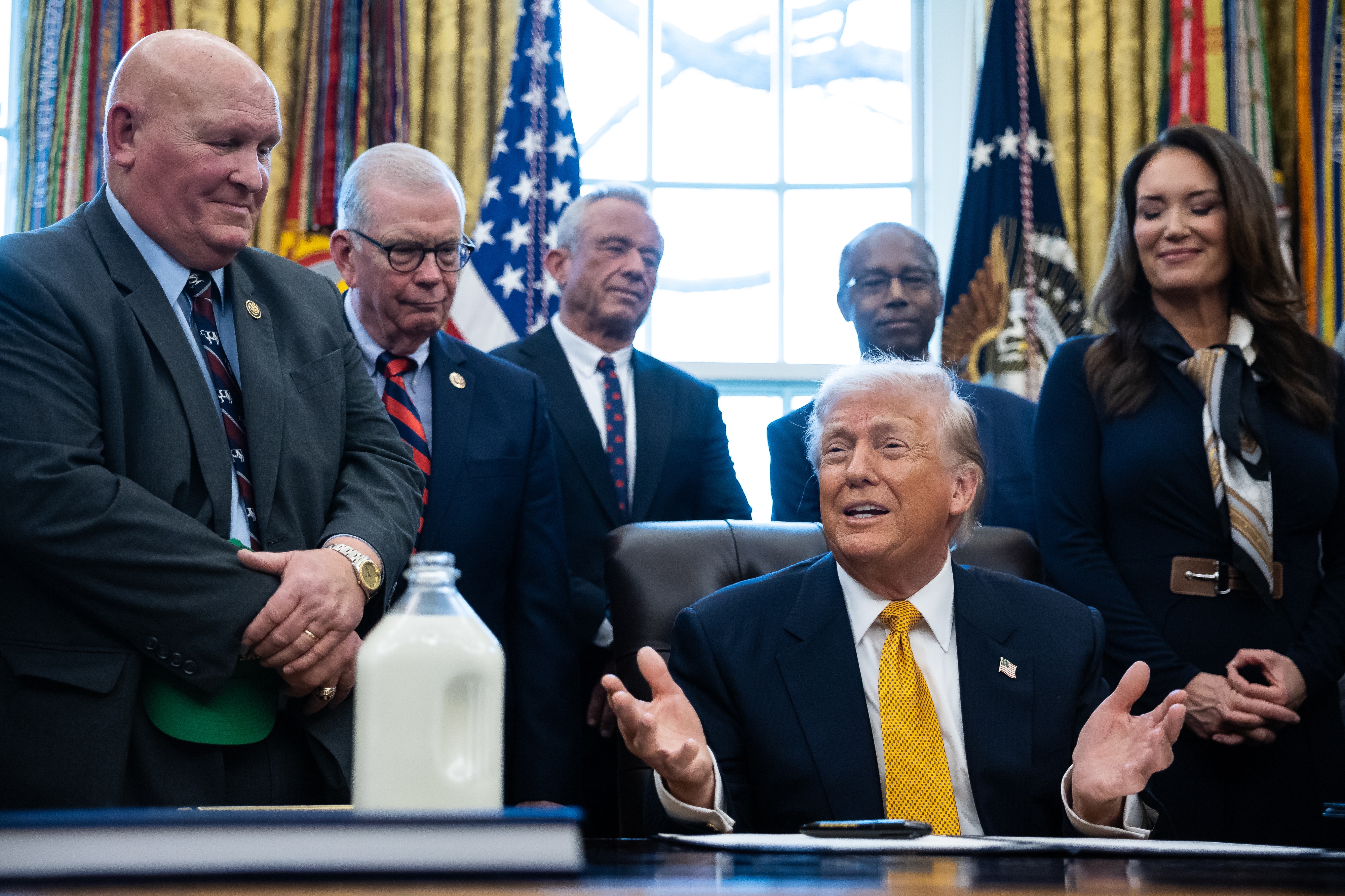 スーツ姿のグループが机の周りに集まり、一人が話している。牛乳瓶が机に置かれている。