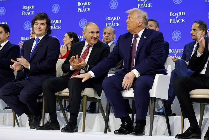 A group of men in suits seated at an event with "Board Peace" written in the background, applauding and conversing