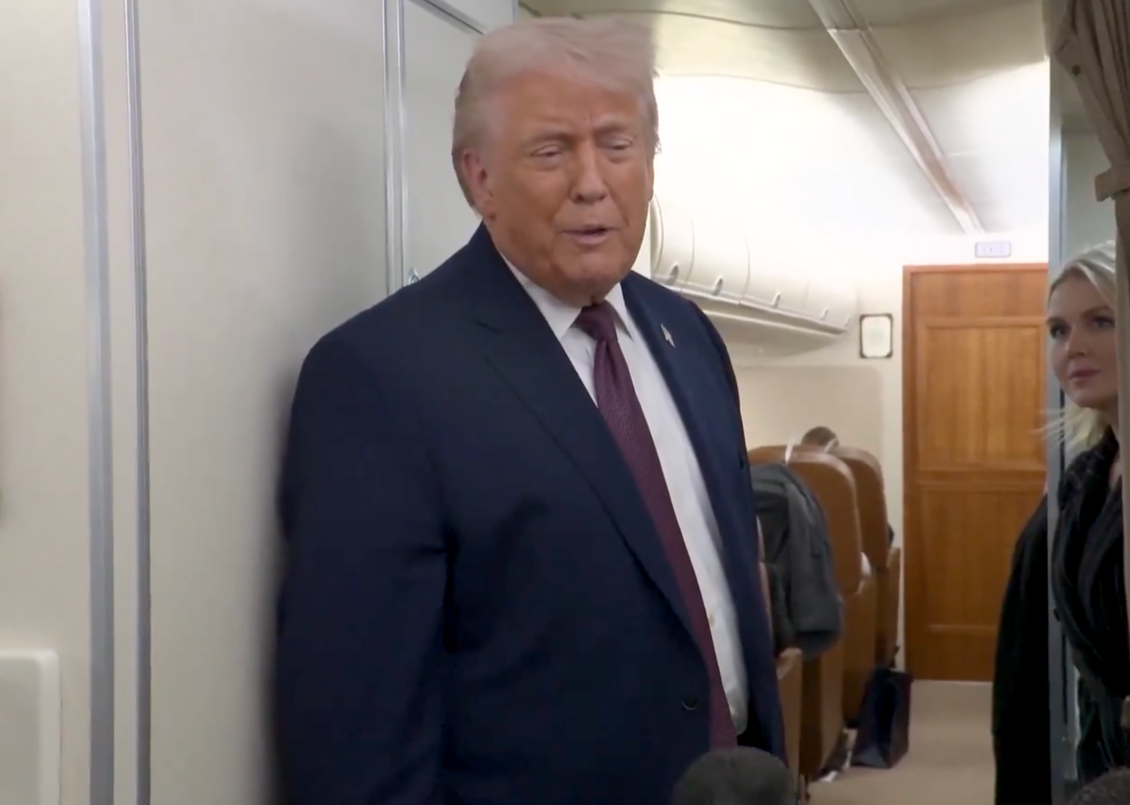 Person in a suit speaking to media on a plane. Passengers and staff are visible in the background